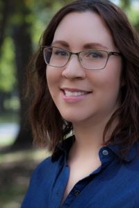 Headshot of Kate with trees in the background. She has brown shoulder length hair, is wearing glasses, a blue shirt, and is smiling.